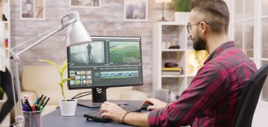 Creative filmmaker working on post production of a movie while working from home. Girlfriend in the background is walking in the house and talking on the phone.