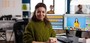 Woman retoucher looking at camera smiling sitting in creative design media agency retouching client photos on PC with two displays. Artist graphic editor working in start up office with digital assets